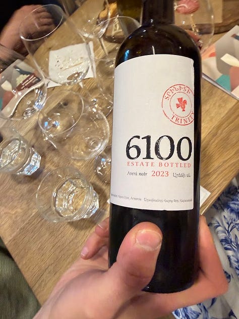 Photos inside In Vino wine bar in Yerevan, including a hand holding a bottle of Trinity Canyon Areni Noir, staff pouring wine behind shelves stocked with Armenian and international bottles, friends toasting with glasses of white wine, a long tasting table set with glassware beneath a sign reading 'The Renaissance of Armenian Wine Making,' and a wall of photos honoring winemakers