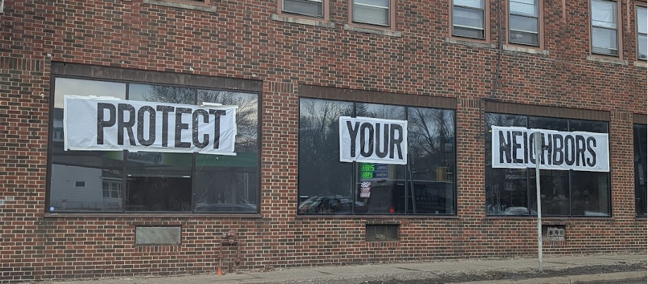 Photograph of a red brick building with three window across the front. In the windows are signs that read protect your neighbors, with one word in each window.