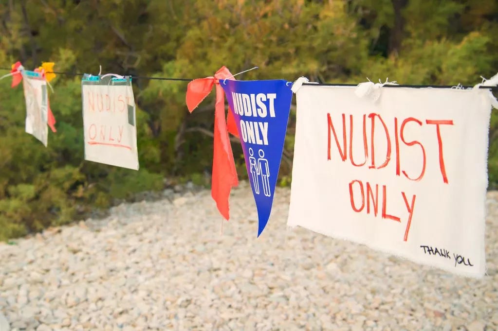 Cloth and plastic signs reading “Nudist Only” hanging on a line near trees and rocks. Cloth and plastic signs reading “Nudist Only” hanging on a line near trees and rocks.