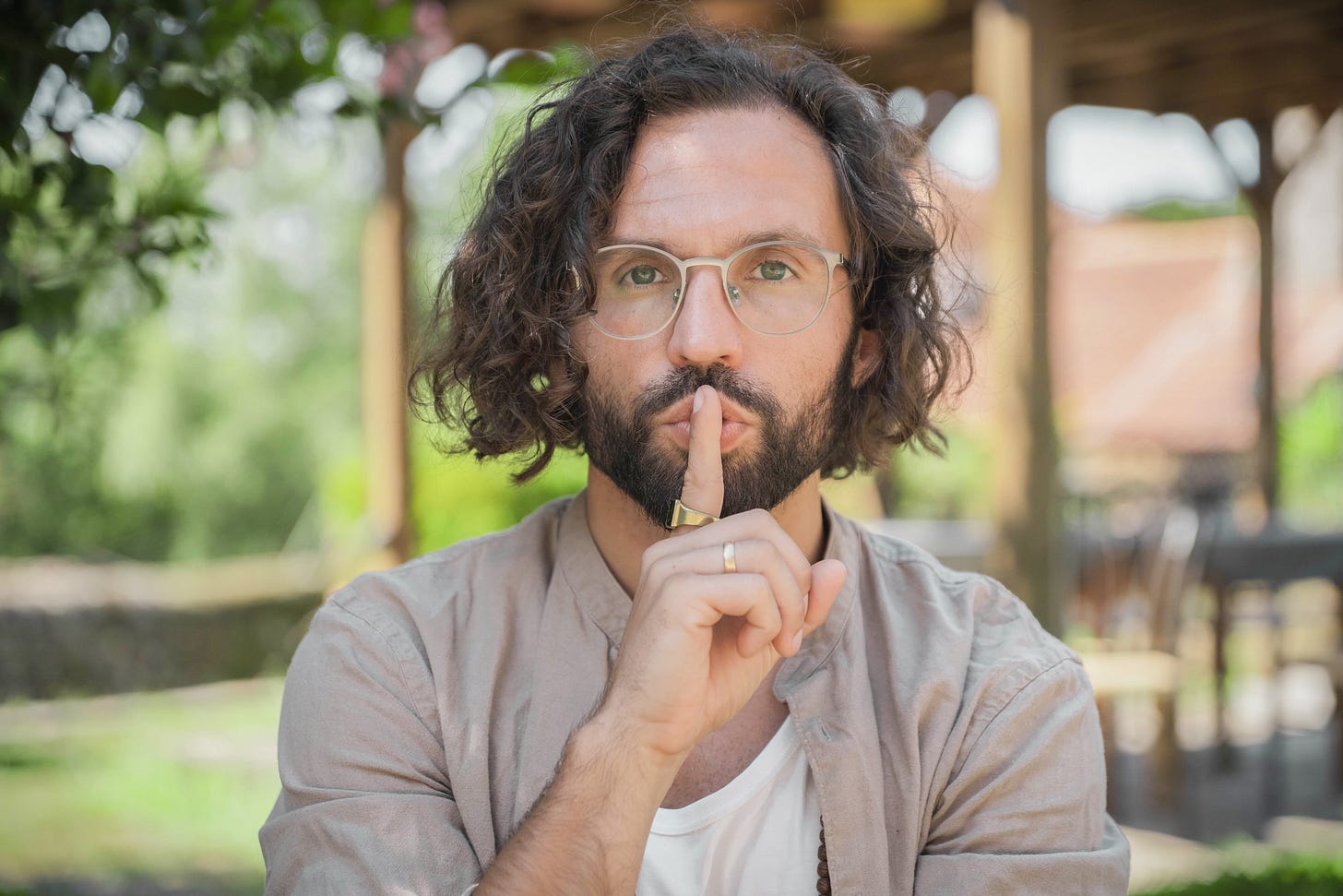 Man with long hair holding a finger to this mouth while standing in a garden