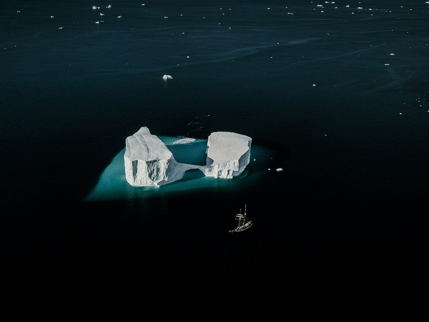 Fotografia de um oceano bem escuro, visto do alto. No centro, é possível ver 2 icebergs separados por um fio de gelo, que dá a impressão de que eles estão segurando as mãos. Ao lado, há um barco. Fotografia de um oceano bem escuro, visto do alto. No centro, é possível ver 2 icebergs separados por um fio de gelo, que dá a impressão de que eles estão segurando as mãos. Ao lado, há um barco.