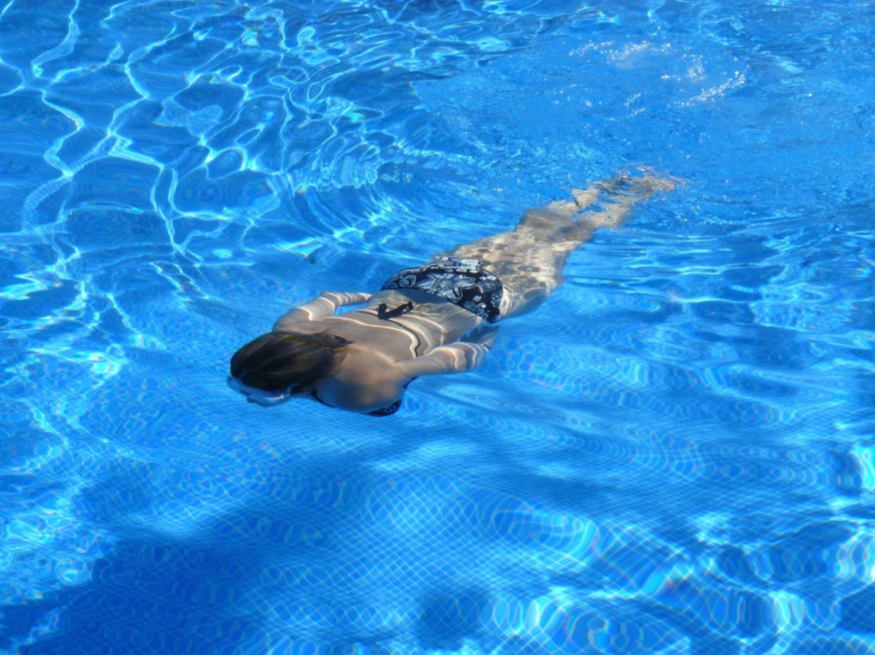 Overhead view of a woman swimming underwater in a bright blue pool, wearing a colorful patterned swimsuit.
