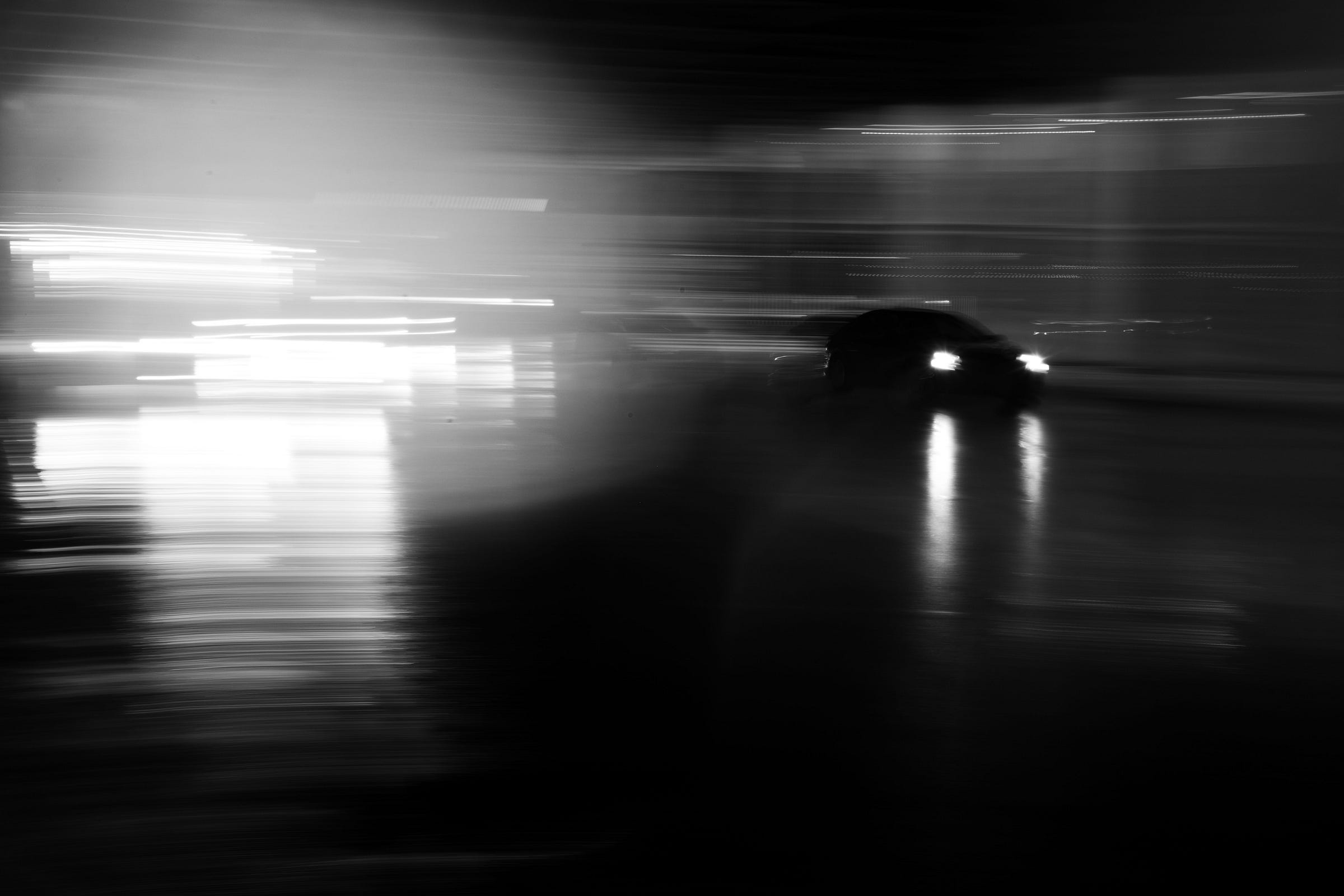 A car with headlights on speeds through a rain-soaked street at night, leaving behind blurred light streaks and reflections on the wet pavement. A car with headlights on speeds through a rain-soaked street at night, leaving behind blurred light streaks and reflections on the wet pavement.