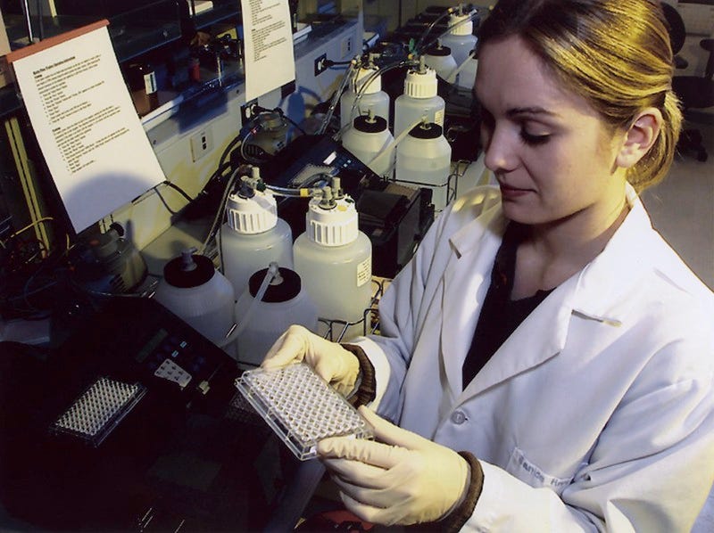 A photo of a researcher in a white lab coat and gloves removing a plate of purified DNA from a plate washer in a lab. A photo of a researcher in a white lab coat and gloves removing a plate of purified DNA from a plate washer in a lab.