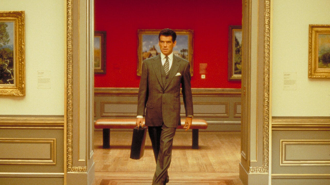 Pierce Brosnan in a natty suit and tie carrying a briefcase walking forward out of a museum room which frames him tastefully