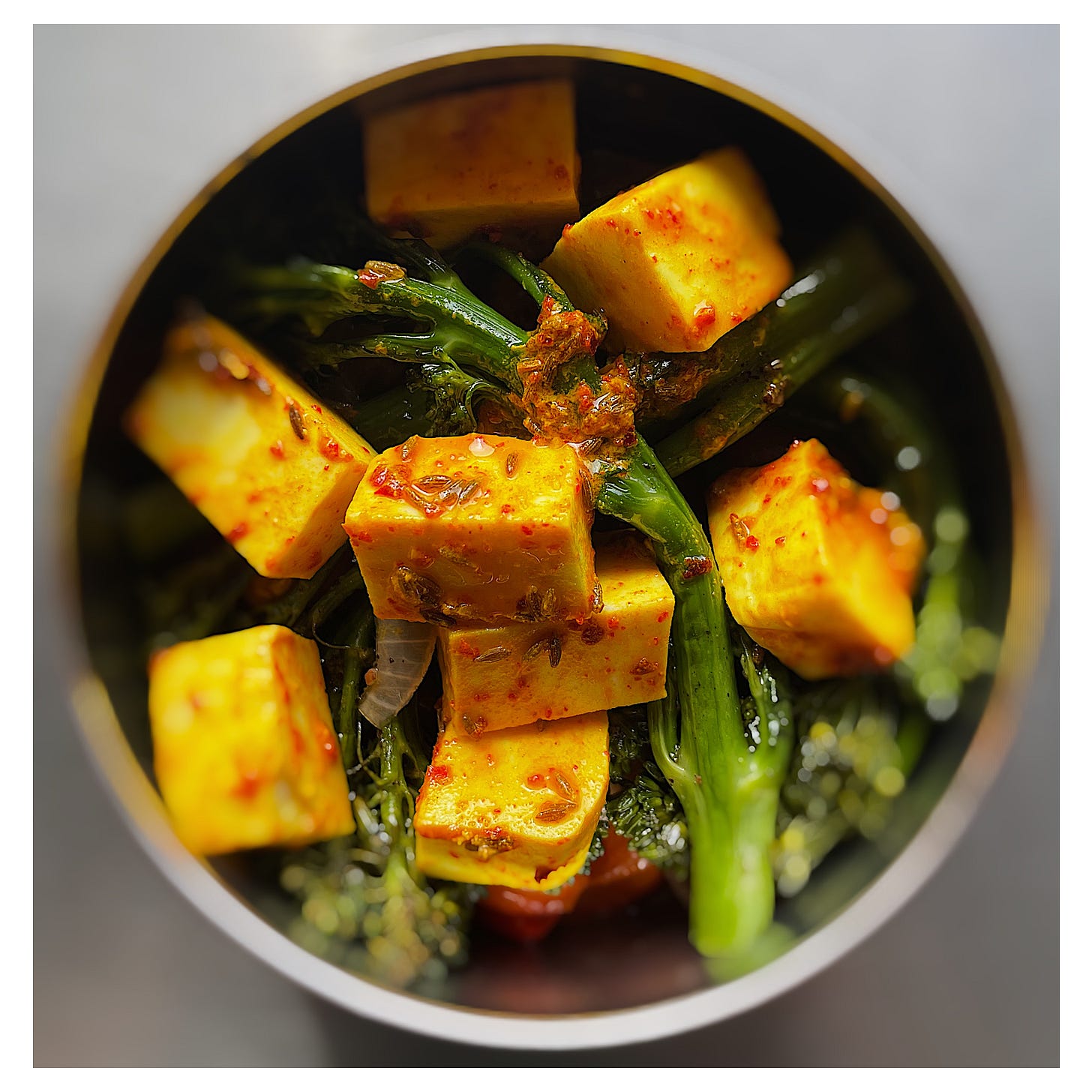 Roast Broccoli, Red Peppers & Paneer in a steel bowl, photographed from above. Most visible are some spears of broccoli and squares of white cheese, coated in yellow oil, cumin seeds and red chilli flakes