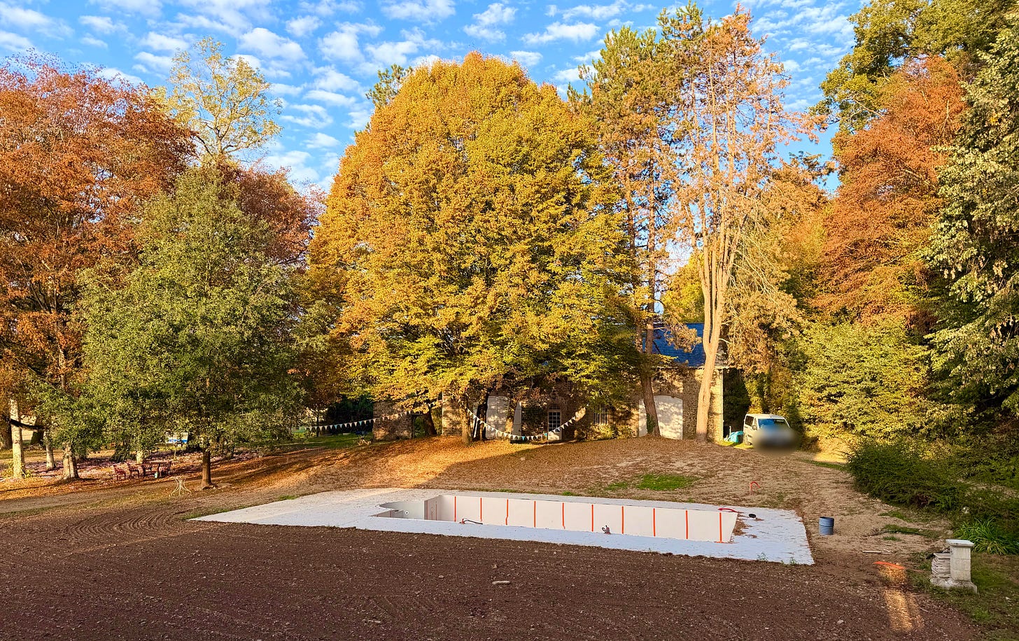 Golden and copper autumn trees on a sunny day behind an old stone coach house hung with bunting, with a rectangular pool under construction in the foreground, its concrete shell edged in white with orange markers, set in freshly turned earth.