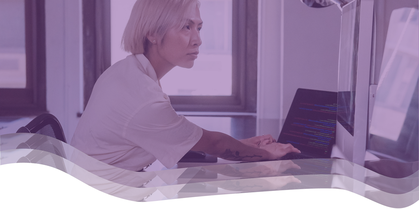 A woman types code on a laptop while glancing at a monitor to her right in an office setting.