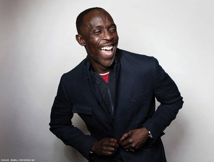 This image shows the actor Michael K. Williams wearing a casual black suit and laughing at something off-camera. This image shows the actor Michael K. Williams wearing a casual black suit and laughing at something off-camera.