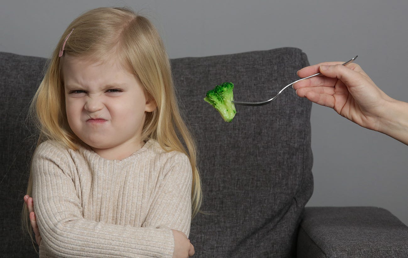 A blonde little girl scrunches up her face and crosses her arms as a adult attempts to feed her broccoli on a fork.