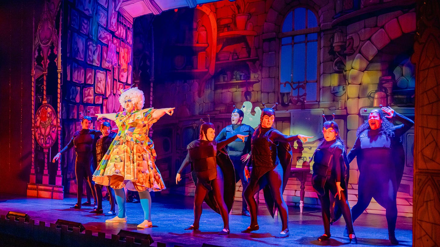 A brightly coloured Dame in a yellow babydoll dress gestures in front of a chorus of black-clad cockroaches.