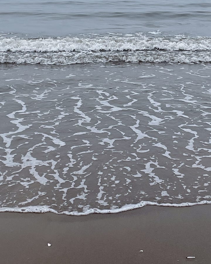Image 1: Holding an almond croissant with a bite taken out, sitting cross-legged. Sand is visible below. / Image 2: The tide rolling in, gentle waves meeting the shore. / Image 3: Inside Tills & Company bookshop—books, tables, and people enjoying a slow moment. / Image 4: A coffee table in Tills with my book Ways of Seeing and a coffee cup resting on it.