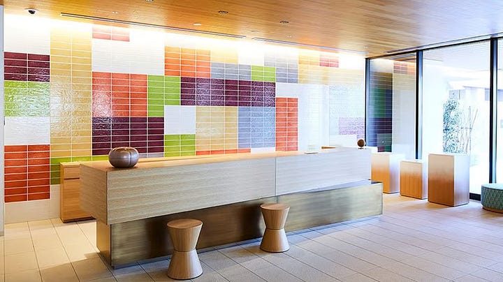 The modern, bright reception area of Hotel Intergate Kanazawa featuring a colorful geometric tiled wall and a long minimalist wooden front desk. An accessible hotel bathroom vanity area with a wide white countertop, a square vessel sink, and open wooden shelving containing towels and a small refrigerator. A spacious accessible hotel room featuring two twin beds, a large brown sofa, and a small round table with colorful patterned chairs and stools. An accessible bathroom featuring a bathtub with a handheld shower head, a sink, and a toilet with grab bars against light wood-textured walls.