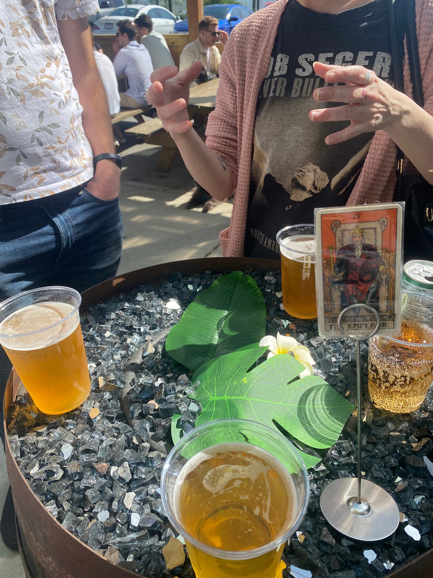 Glasses of beer on top of a barrel covered in dark shiny rocks, and a couple of fake tropical leaves and flowers. Behind it Jesse and Steph are standing, not fully visible. Steph is wearing a Bob Seger shirt and gesturing with both hands. In the foreground a laminated tarot card, 'the emperor', is on a stand, indicating we're waiting for food to be brought to the table..