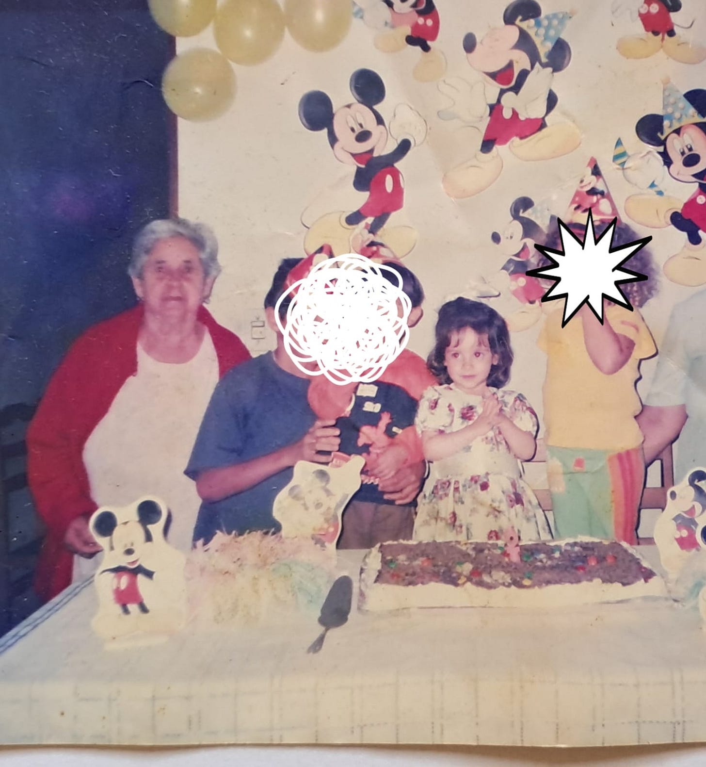 A foto, com aspecto antigo e levemente desbotado, mostra um grupo de pessoas em volta de uma mesa, com decorações do Mickey Mouse penduradas na parede. No centro da cena, há dois bolos de aniversário e uma espátula, sugerindo uma festa.  À esquerda, uma senhora de cabelos curtos, grisalhos e cacheados, com uma blusa branca e um casaco de lã vermelho, sorri levemente. Ao seu lado, está uma criança com blusa azul e um menino menor, de blusa vermelha.  No lado direito, há uma menina com um vestido floral, que está com as mãos juntas, olhando para a câmera. Ao lado dela, há outra criança, parcialmente visível. Todos parecem felizes, celebrando o aniversário.