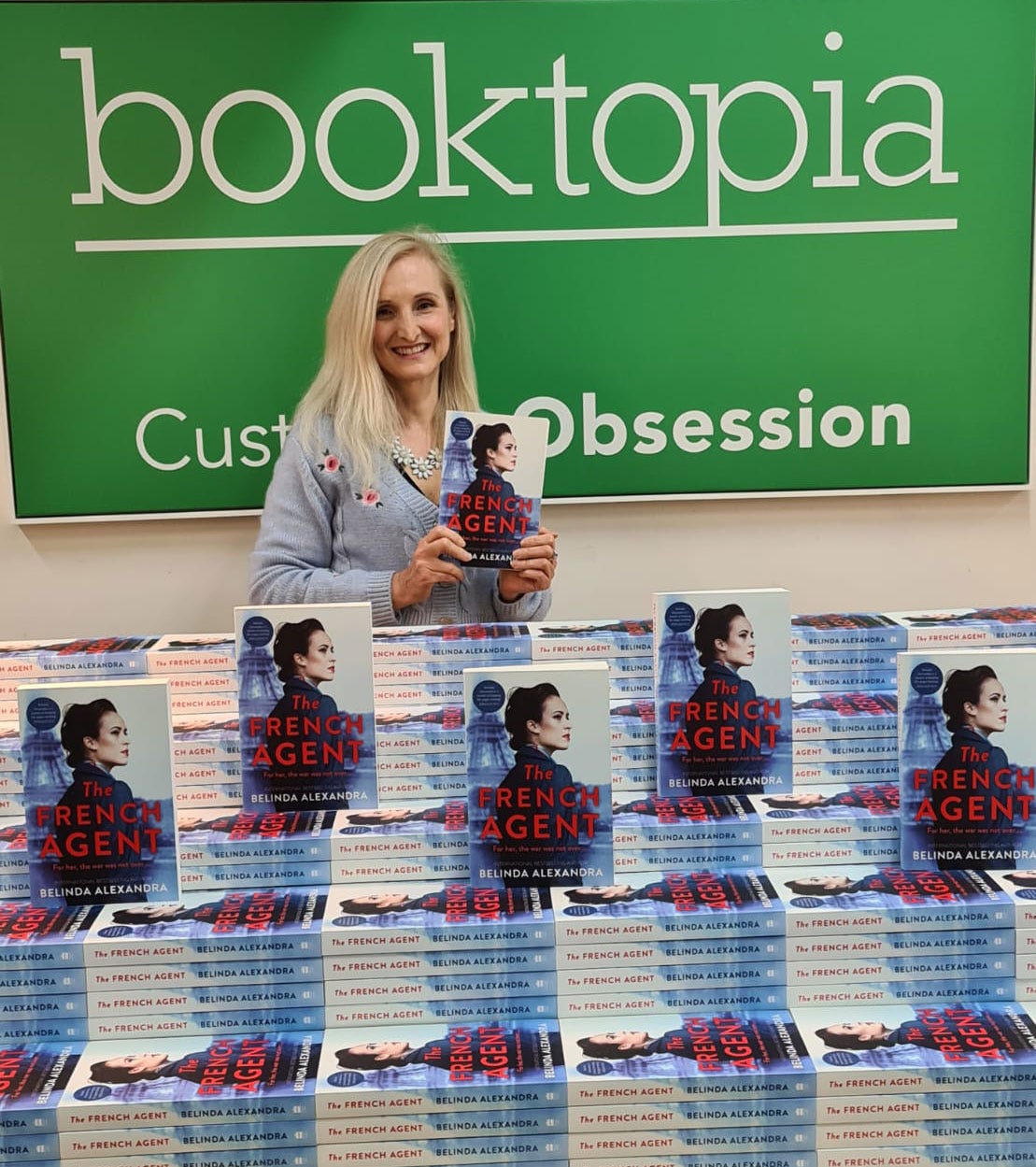 Green banner in the back: Booktopia. Custom Obsession. Standing in front is Belinda, long blond hair smiling holding a copy of The French Agent. In front of her a table with mountains of copies of The French Agent.  