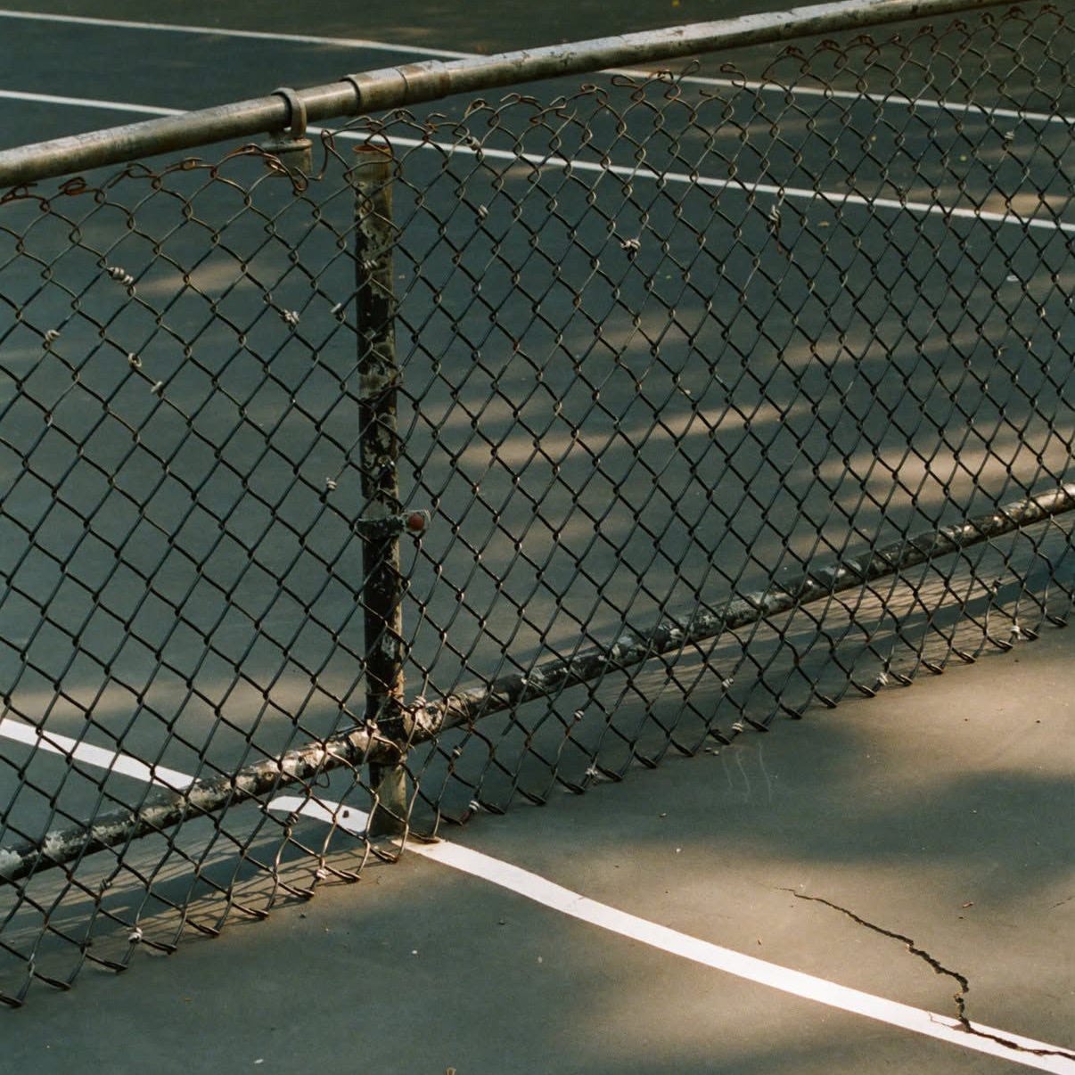 A worn public tennis court at Marcy Playground in Brooklyn, with a weathered chain-link fence and faded green court surface. A worn public tennis court at Marcy Playground in Brooklyn, with a weathered chain-link fence and faded green court surface.