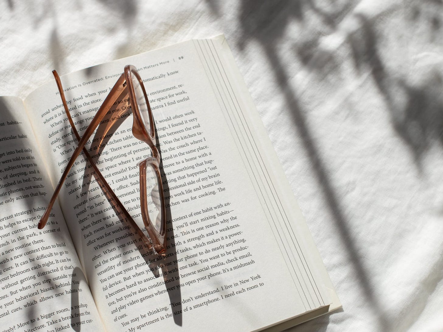 A pair of brown reading glasses resting on an open book, dappled in soft sunlight and shadows from nearby leaves. The scene suggests calm, reflection, and quiet reading moments.