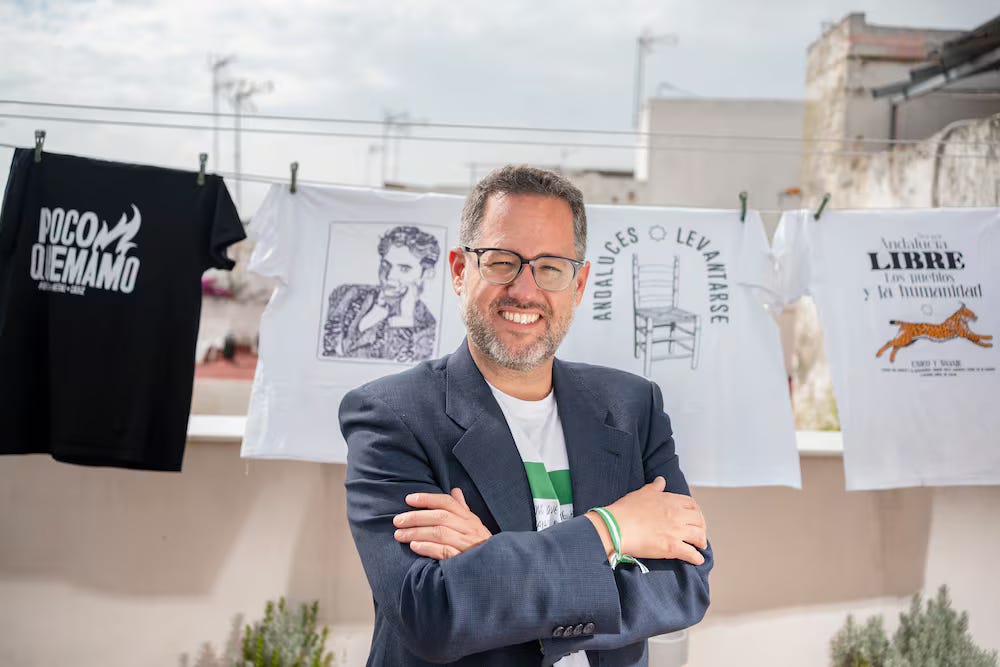 Un uomo sorridente con gli occhiali e la barba corta posa a braccia conserte su una terrazza, davanti a un filo steso con diverse magliette stampate con scritte e grafiche in spagnolo. Un uomo sorridente con gli occhiali e la barba corta posa a braccia conserte su una terrazza, davanti a un filo steso con diverse magliette stampate con scritte e grafiche in spagnolo.