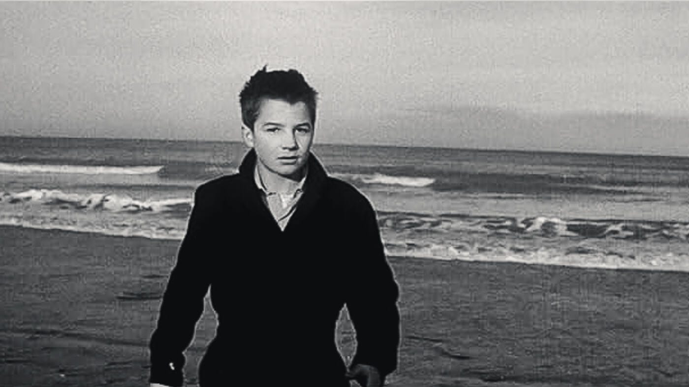 Antoine Doinel stands alone on a beach facing the camera, with the ocean waves behind him in the iconic final shot of the film.
