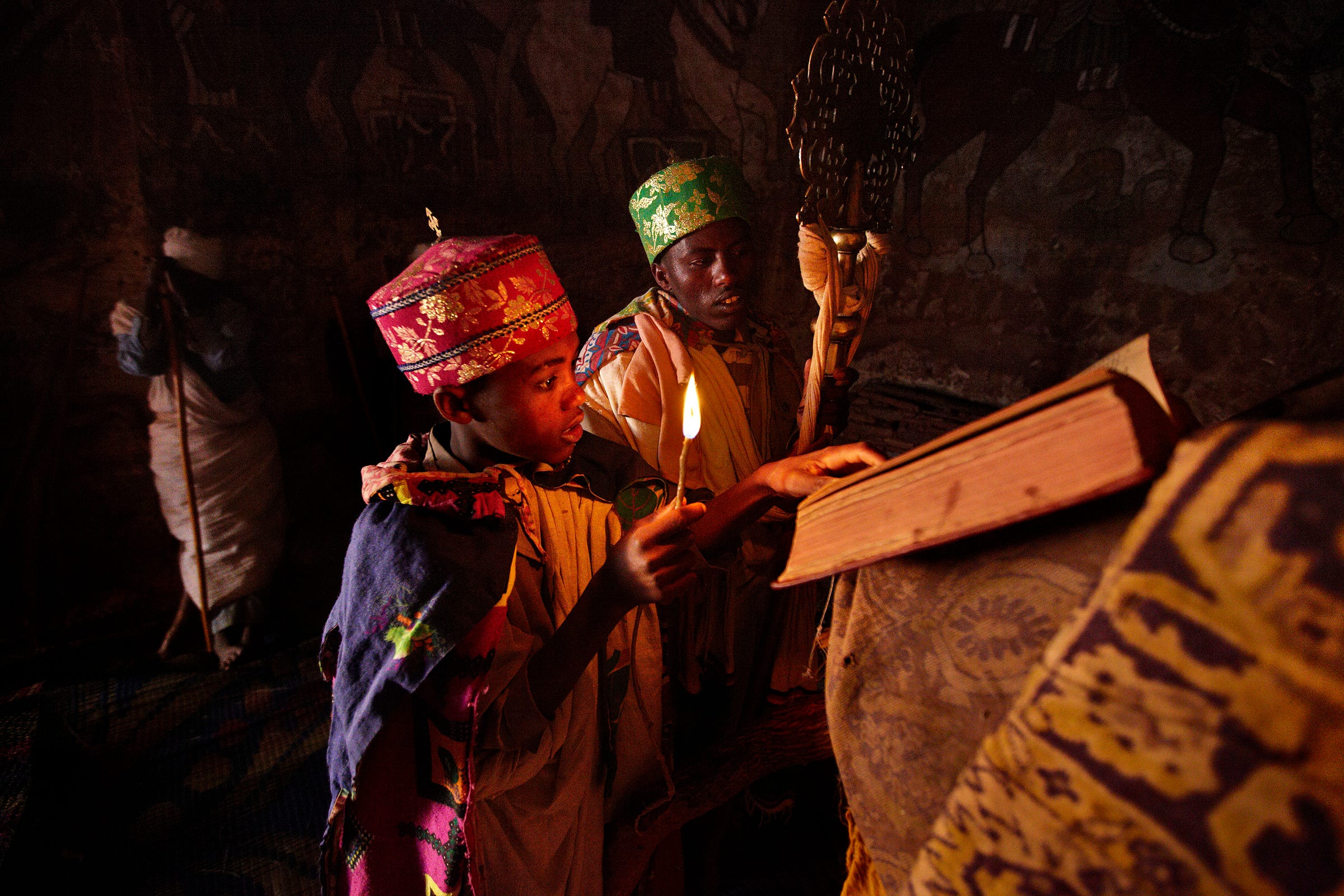 A young boy in colorful traditional attire holds a candle and reads from an ancient manuscript, standing beside another person in ceremonial clothing, inside a dimly lit, ornately decorated room. A young boy in colorful traditional attire holds a candle and reads from an ancient manuscript, standing beside another person in ceremonial clothing, inside a dimly lit, ornately decorated room.