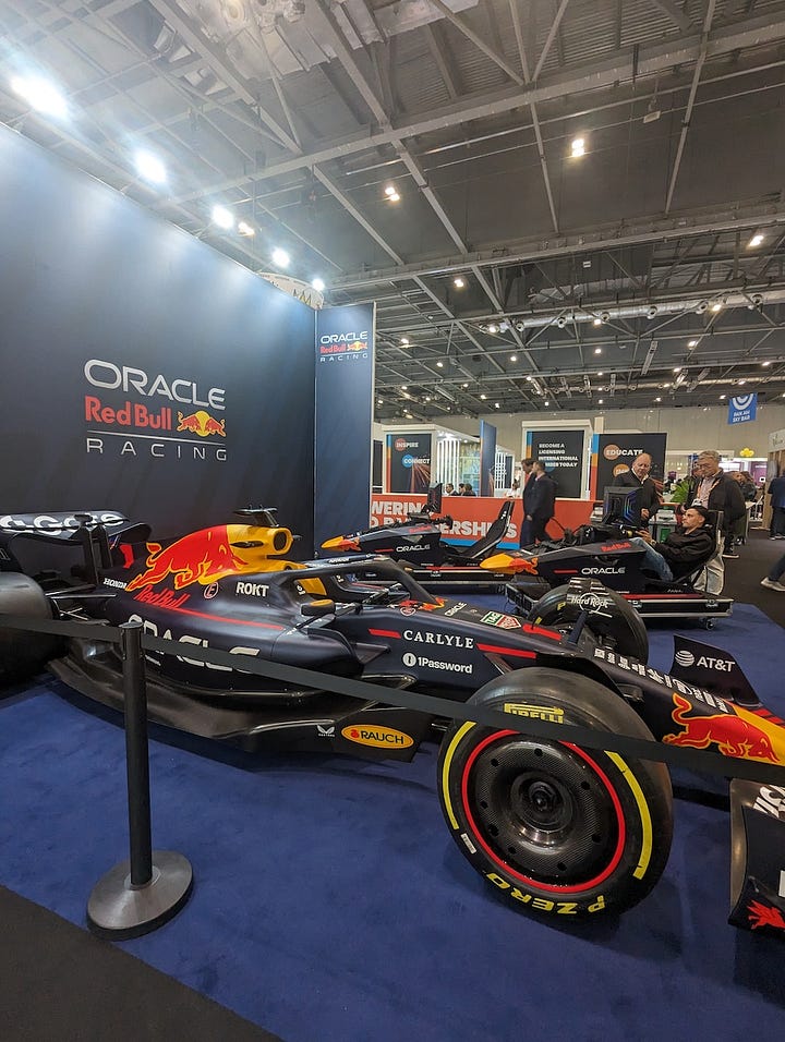 A trade show scene featuring two distinct exhibits: one showcasing an Oracle Red Bull Racing Formula 1 car displayed against a branded backdrop with sponsors such as Oracle, Red Bull, and Pirelli visible under bright convention hall lighting; and another booth promoting the Skibidi brand, where mannequins dressed in Gen Alpha–style streetwear stand beside shelves filled with Skibidi merchandise and Funko Pop figures, illuminated by striking red neon lights as visitors browse in the background.