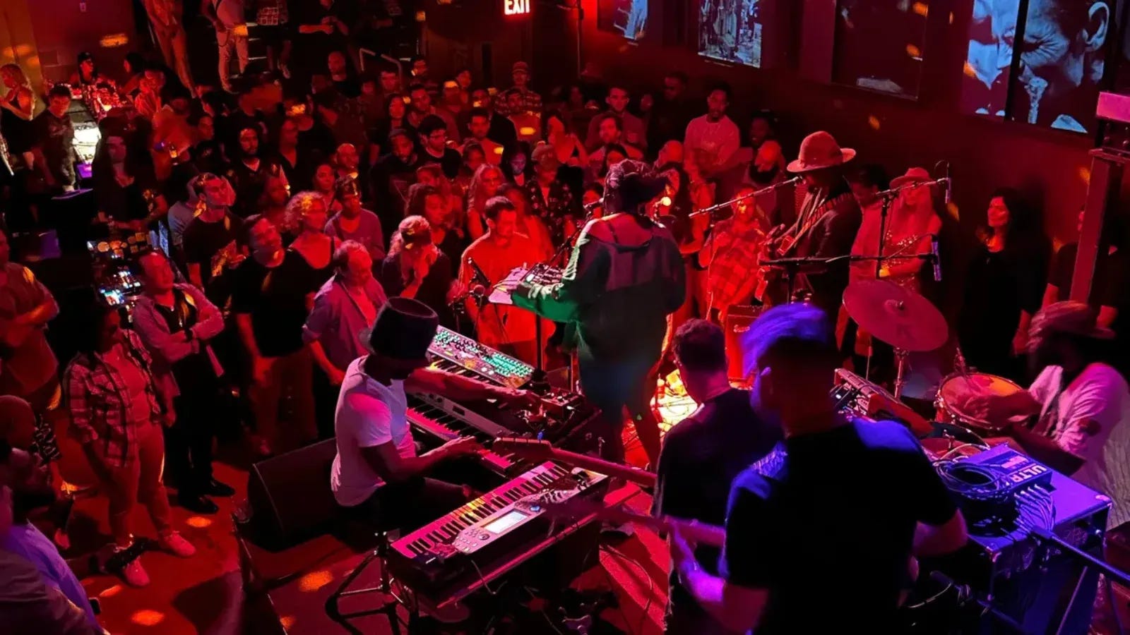 Producer Mondays from above at Nublu, the room washed red: a keyboard player in a flat-brim hat at stacked keys, a singer at the mic, a guitarist in a wide-brim hat, and the crowd packed right up to the players
