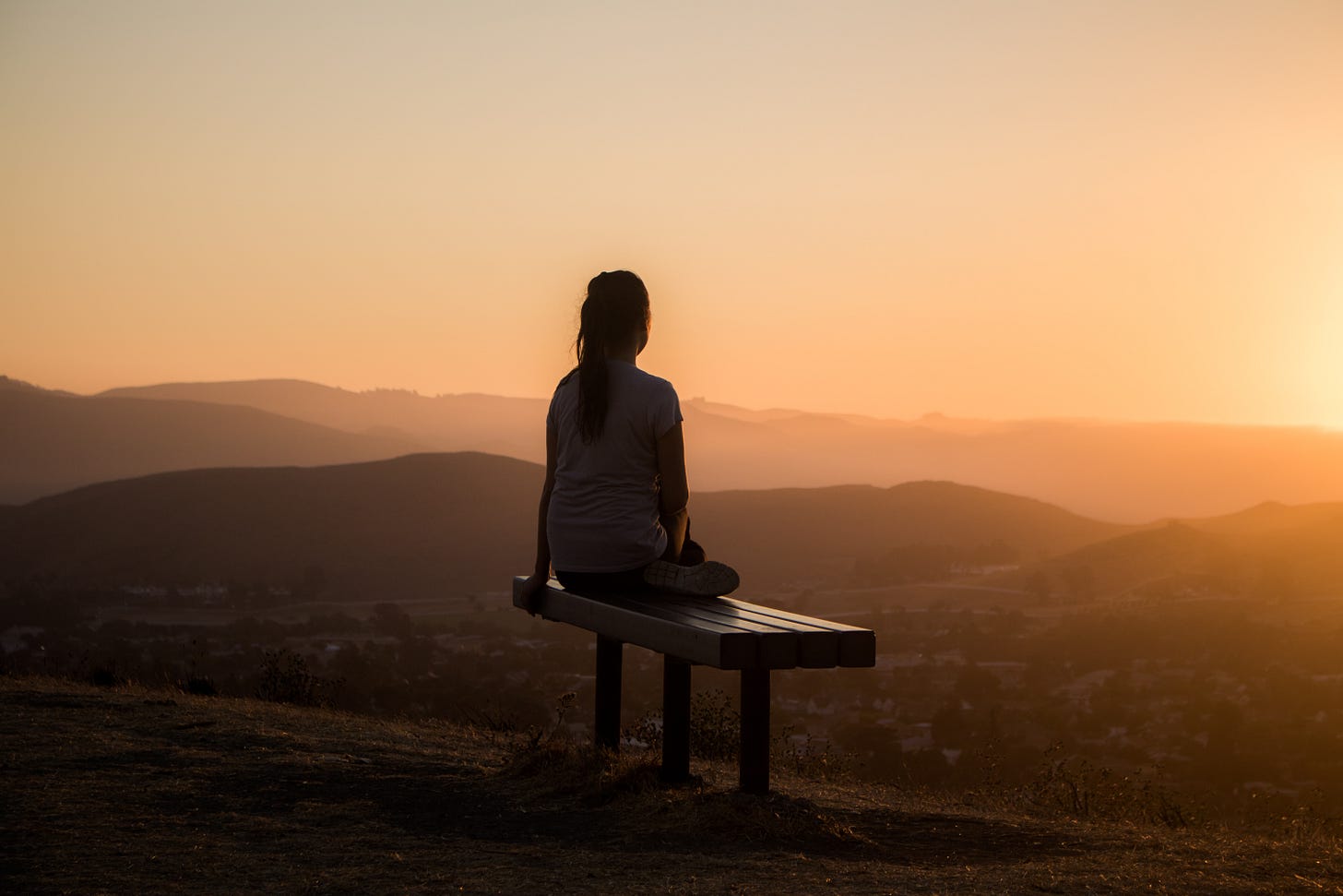 A woman sitting on a bench looking out to the right. Her back is to the camera. Her silhouette contrasts against a peach-colored sunrise. A woman sitting on a bench looking out to the right. Her back is to the camera. Her silhouette contrasts against a peach-colored sunrise.