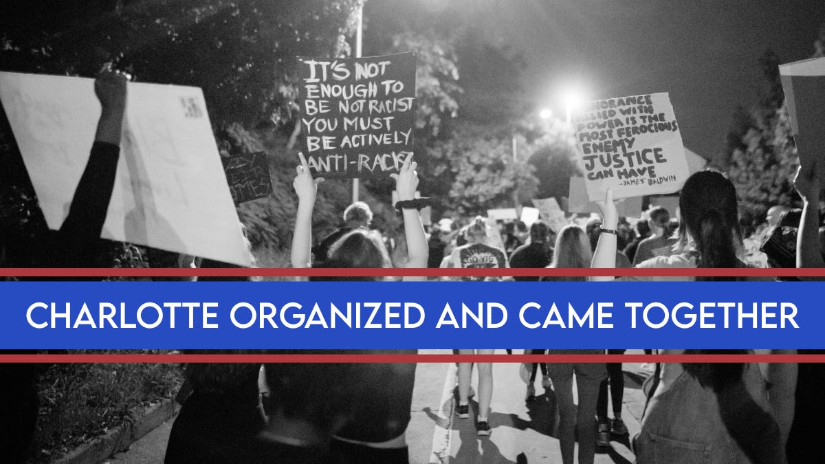 Black-and-white photo of a nighttime protest in Charlotte with a crowd marching and holding signs. Visible signs read ‘It’s not enough to be not racist, you must be actively anti-racist’ and a James Baldwin quote about justice. A blue banner across the image says ‘Charlotte organized and came together.’