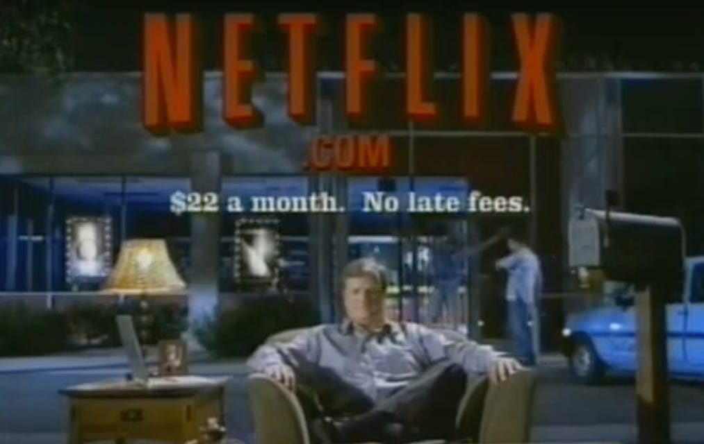Screenshot of a vintage Netflix commercial with a man sitting on a sofa next to a mailbox and the text at the top: Netflix.com $22 a month no late fees.