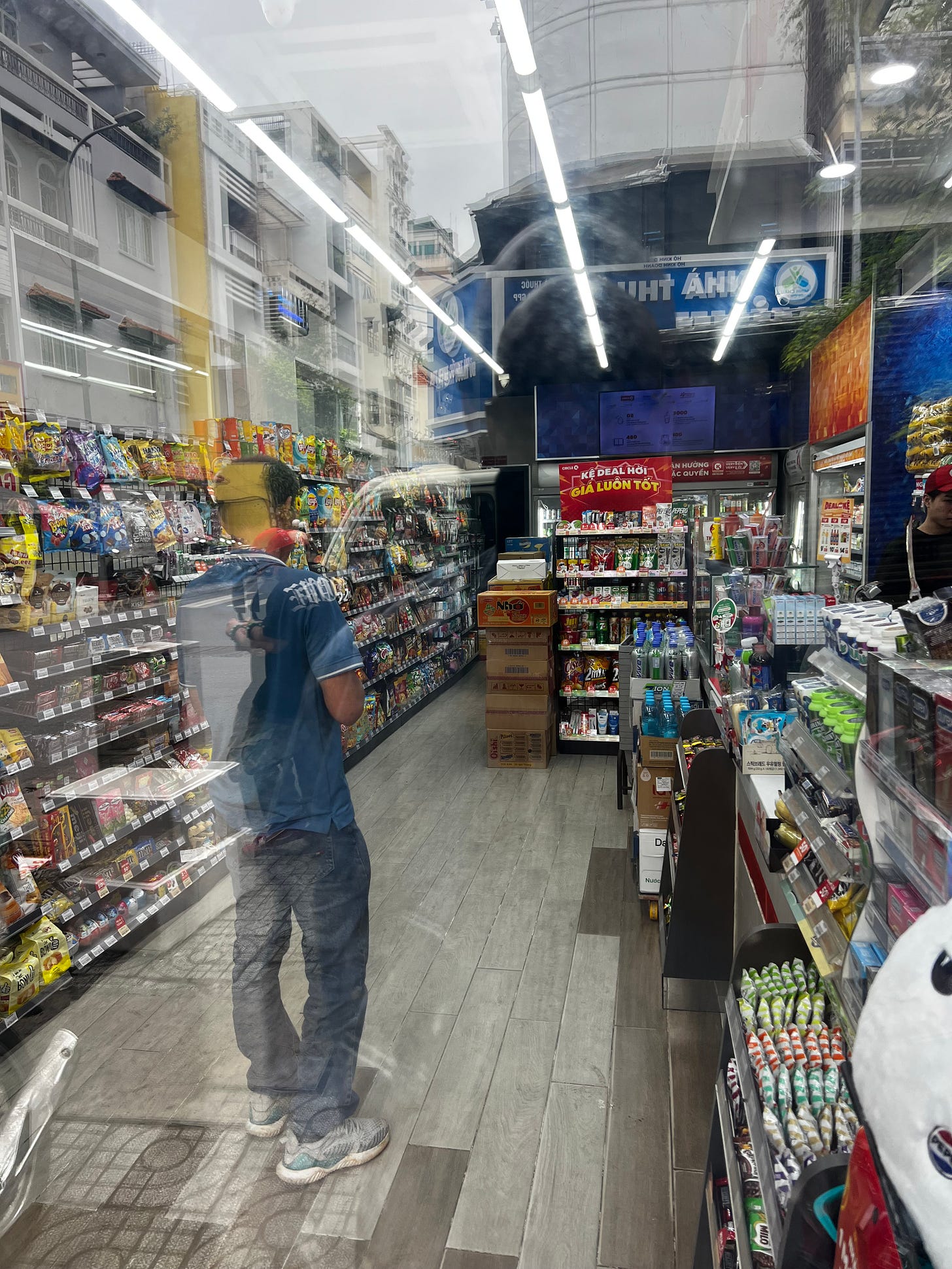 Interior of a Vietnamese convenience store seen through glass, with reflections of the street visible in the window. Shelves of snacks and drinks line the walls, and a customer in a blue shirt stands in the aisle. Outside, motorcycles and buildings are visible in the reflection. Interior of a Vietnamese convenience store seen through glass, with reflections of the street visible in the window. Shelves of snacks and drinks line the walls, and a customer in a blue shirt stands in the aisle. Outside, motorcycles and buildings are visible in the reflection.