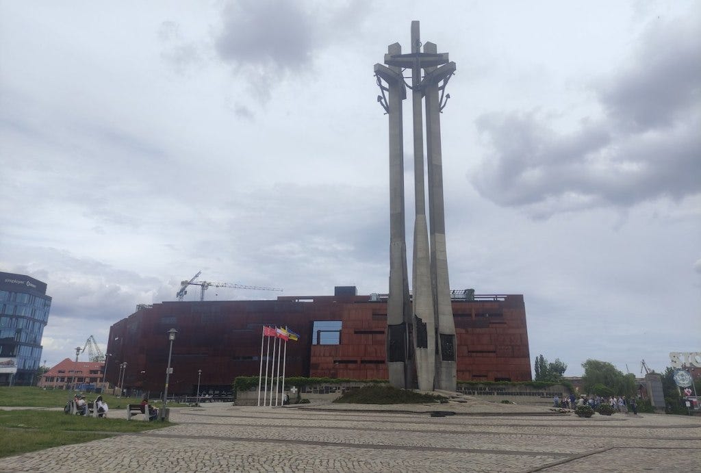 Tall statue, with three concrete legs with crosses at the top, supporting large anchors in memorial to the steel workers killed in 1970. Rusty-looking Solidarity museum is behind; it is a multi-storey building, but nowhere near as high as the statue.