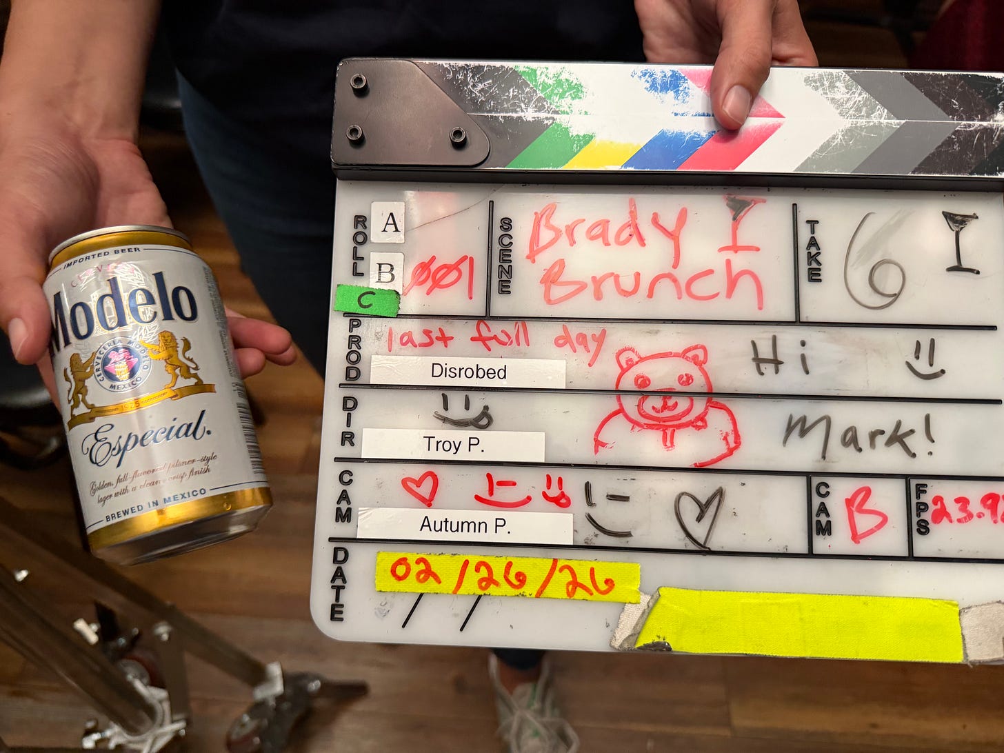 A close-up of a film slate labeled “Disrobed” covered in handwritten notes, doodles, and signatures, including “Brady Brunch,” “last full day,” and small drawings. A person holds the slate while another hand holds a can of Modelo beer beside it, suggesting the end of a long shoot day.