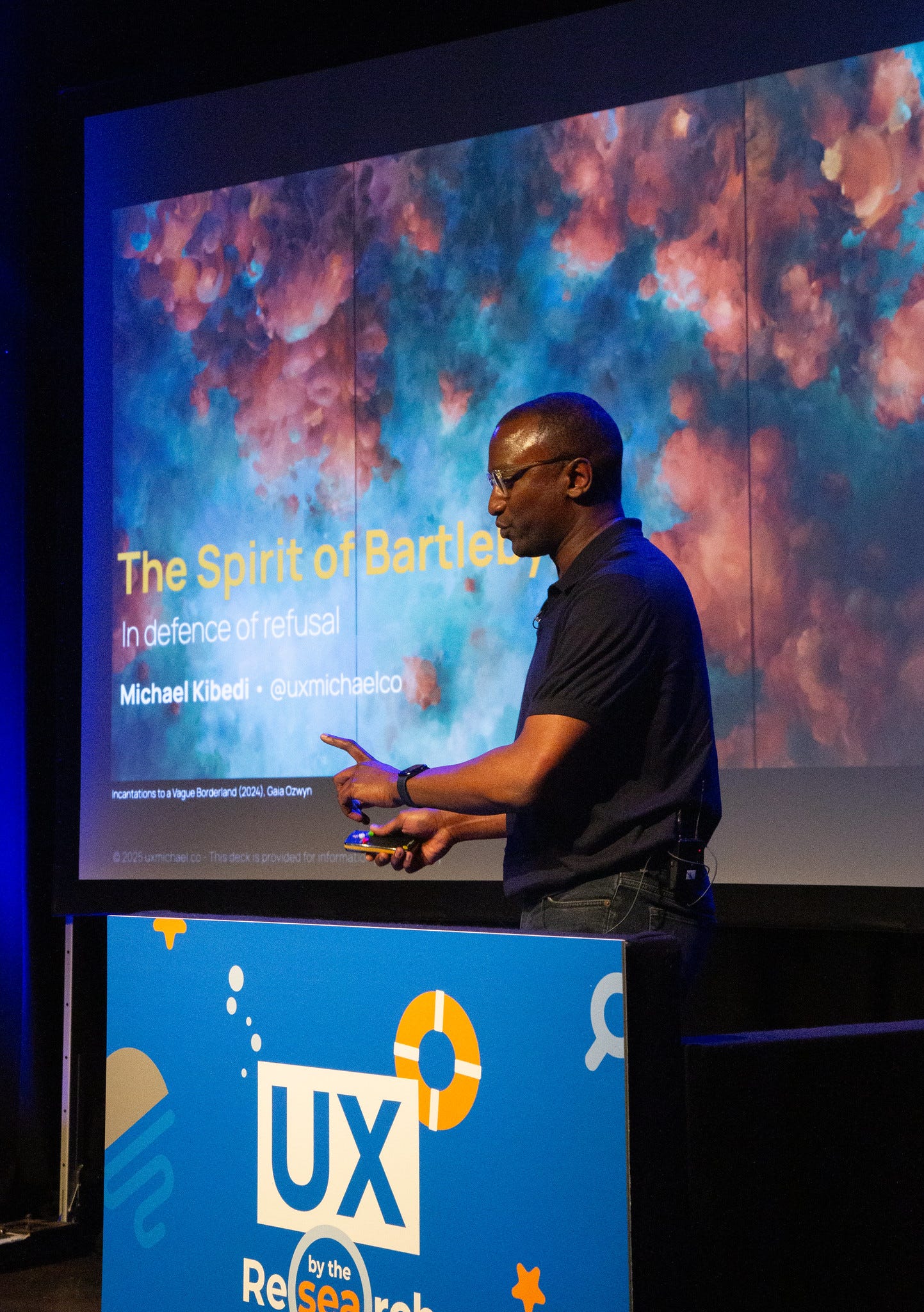 Photo of Michael Kibedi, a tall Black man, giving a talk onstage in a dimly lit auditorium. Photo of Michael Kibedi, a tall Black man, giving a talk onstage in a dimly lit auditorium.