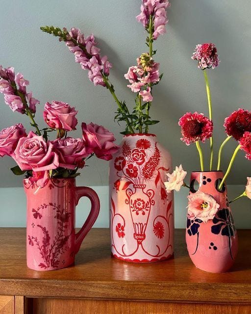 This may contain: three pink vases with flowers in them on a table next to a wall and a wooden cabinet This may contain: three pink vases with flowers in them on a table next to a wall and a wooden cabinet