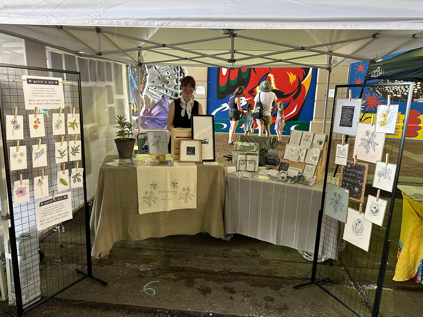 A photo of my artist booth at the festival. I'm a white woman standing behind the booth, which has two tables and two standing grids where art is displayed.