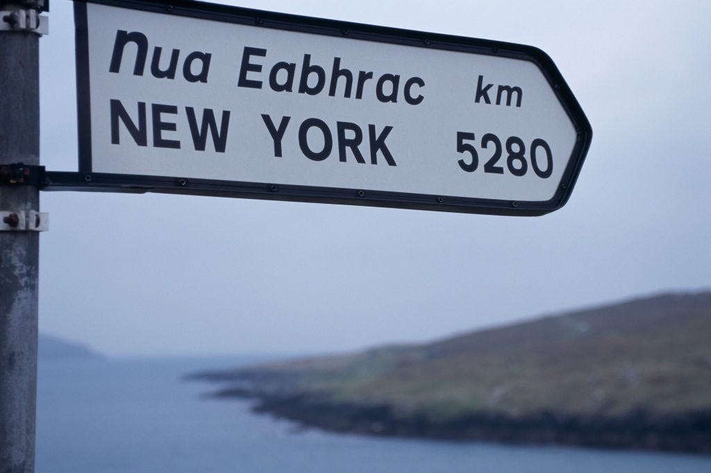 Road sign in Irish and English points to New York, 5280 kilometers away, with a coastal landscape in the background.