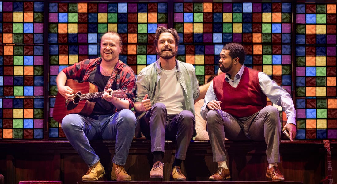A group of three men sitting on stage with a colourful glass background behind them A group of three men sitting on stage with a colourful glass background behind them
