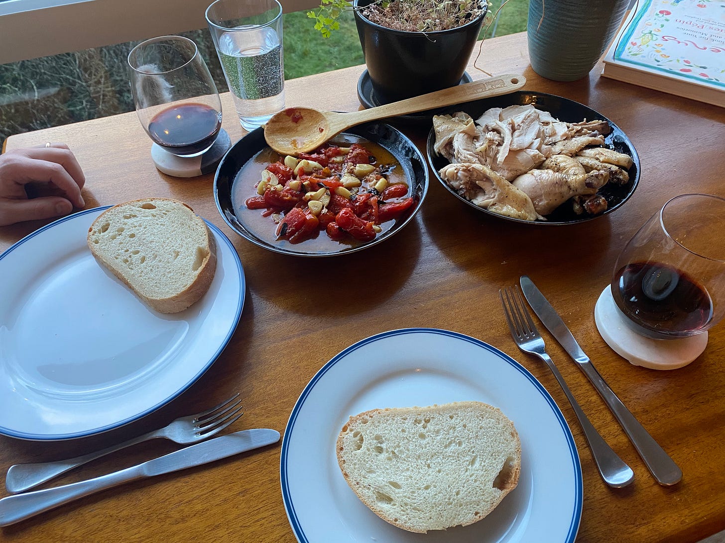 A wood dining table set with plates, cutlery, glasses of red wine on coasters. On each plate is a slice of country bread. Two black serving dishes are in the middle of the table, one with a saucy mix of grape tomatoes, herbs, and roasted garlic, the other with carved chicken pieces. There's a wooden spoon in the tomato dish.