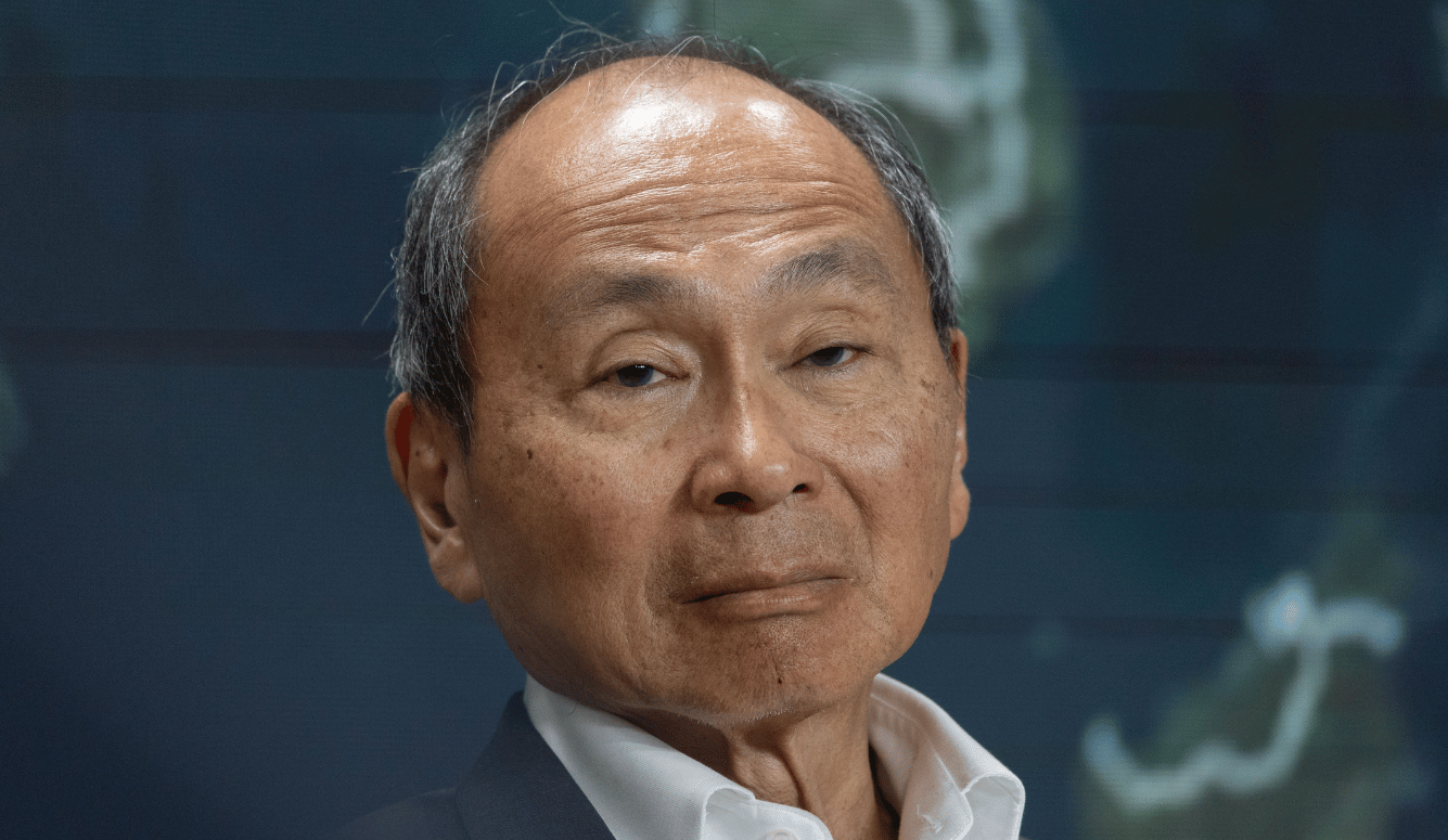 Close-up of Francis Fukuyama at the YES forum in Kyiv, wearing a dark suit and white shirt, with a serious expression and a blurred map in the background.