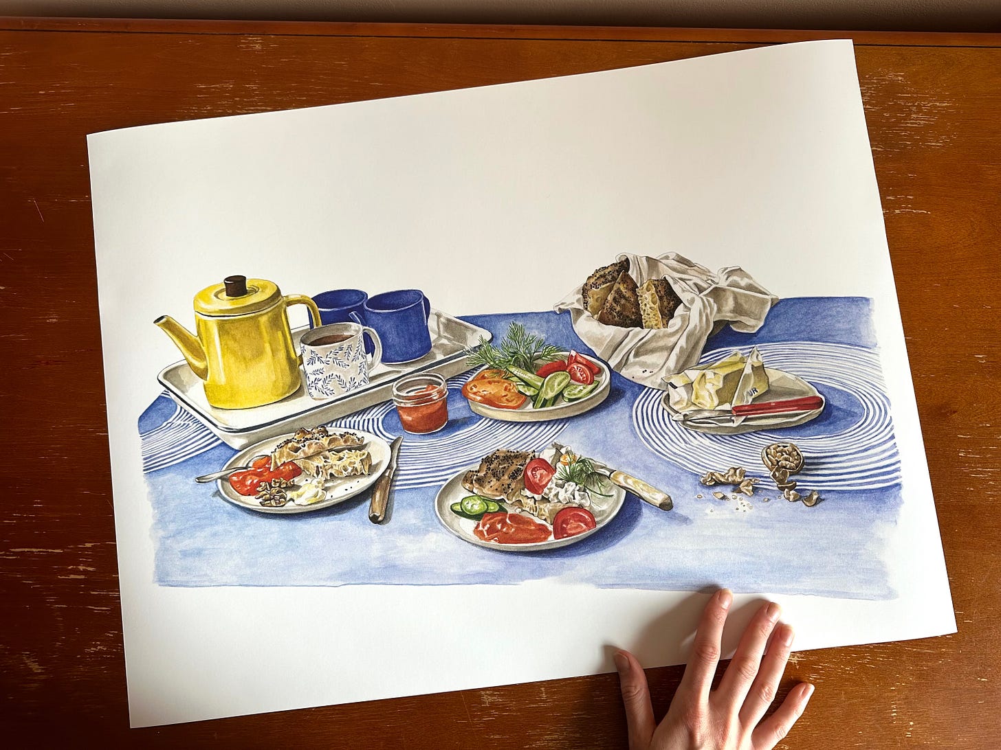 A painting of a blue and white tablecloth with blue mugs and a bright yellow tea pot on a tray, with a couple of plates filled with focaccia, apricot jam, walnuts, tomato slices and cucumber slices. over to the right top corner of the painting is a basket filled with more focaccia with little nigella and sesame seeds dotting it. A little jar of jam and a plate of butter sit on the tablecloth as well. 