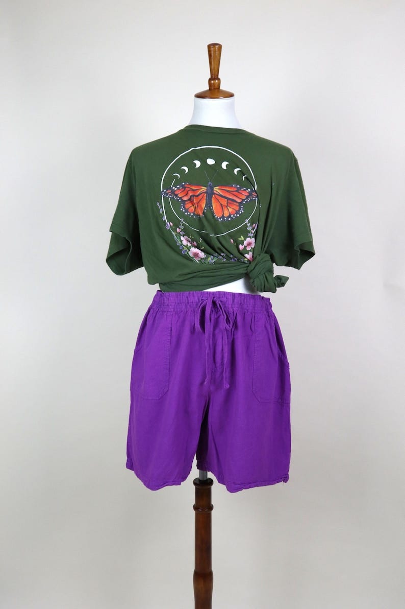May include: An olive green t-shirt with a butterfly graphic and purple shorts. The t-shirt features a circular design with a butterfly and floral accents. The shorts are a vibrant purple with a drawstring waist.