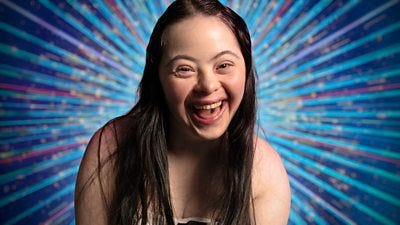 A head and shoulders shot of Ellie Goldstein, a young woman with long dark hair, smiling for the camera against a glittering blue background