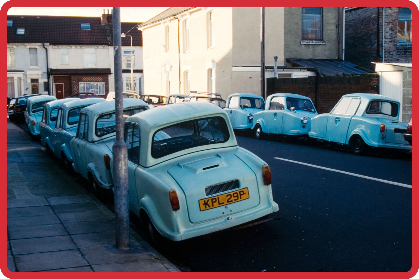 A vintage photo of around a dozen blue invalid carriages parked on a residential English street. The carriages are mostly facing away from us, so we see their car boots and numberplates. A vintage photo of around a dozen blue invalid carriages parked on a residential English street. The carriages are mostly facing away from us, so we see their car boots and numberplates.
