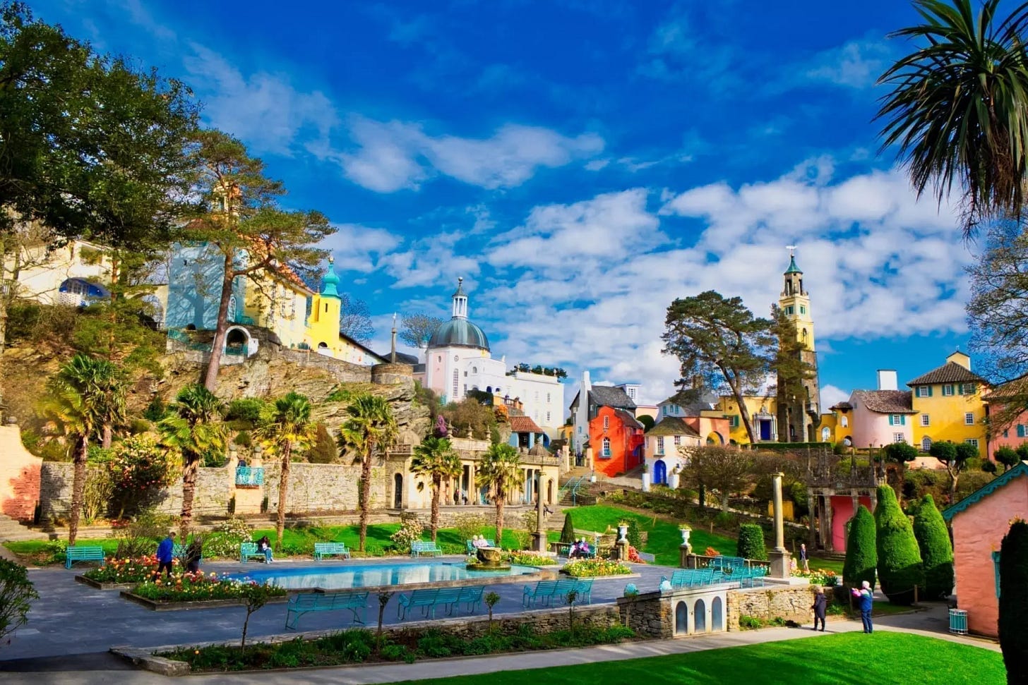 A bright, colorful daytime photograph of the iconic Italianate village of Portmeirion in Gwynedd, North Wales. The scene features pastel-colored towers and houses nestled on a lush hillside, overlooking manicured green lawns, vibrant gardens, palm trees, and a central ornamental pool under a clear blue sky.