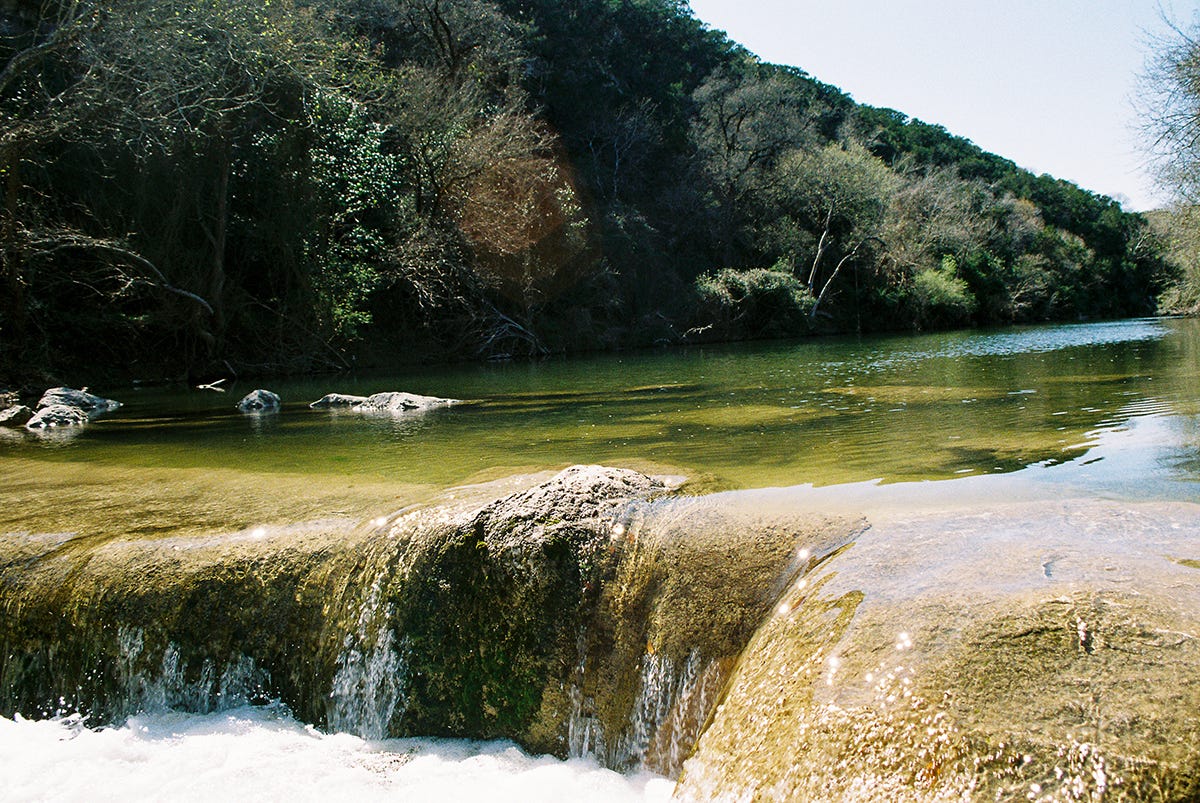 The river water, clear and pale green, cuts the image in half, lens flares sparkle below on the waterfall while a lens flare brings the implication of sun to the dense, green forest on the hill that rises up into the horizon. A layer of thin white foam splashes at the bottom.