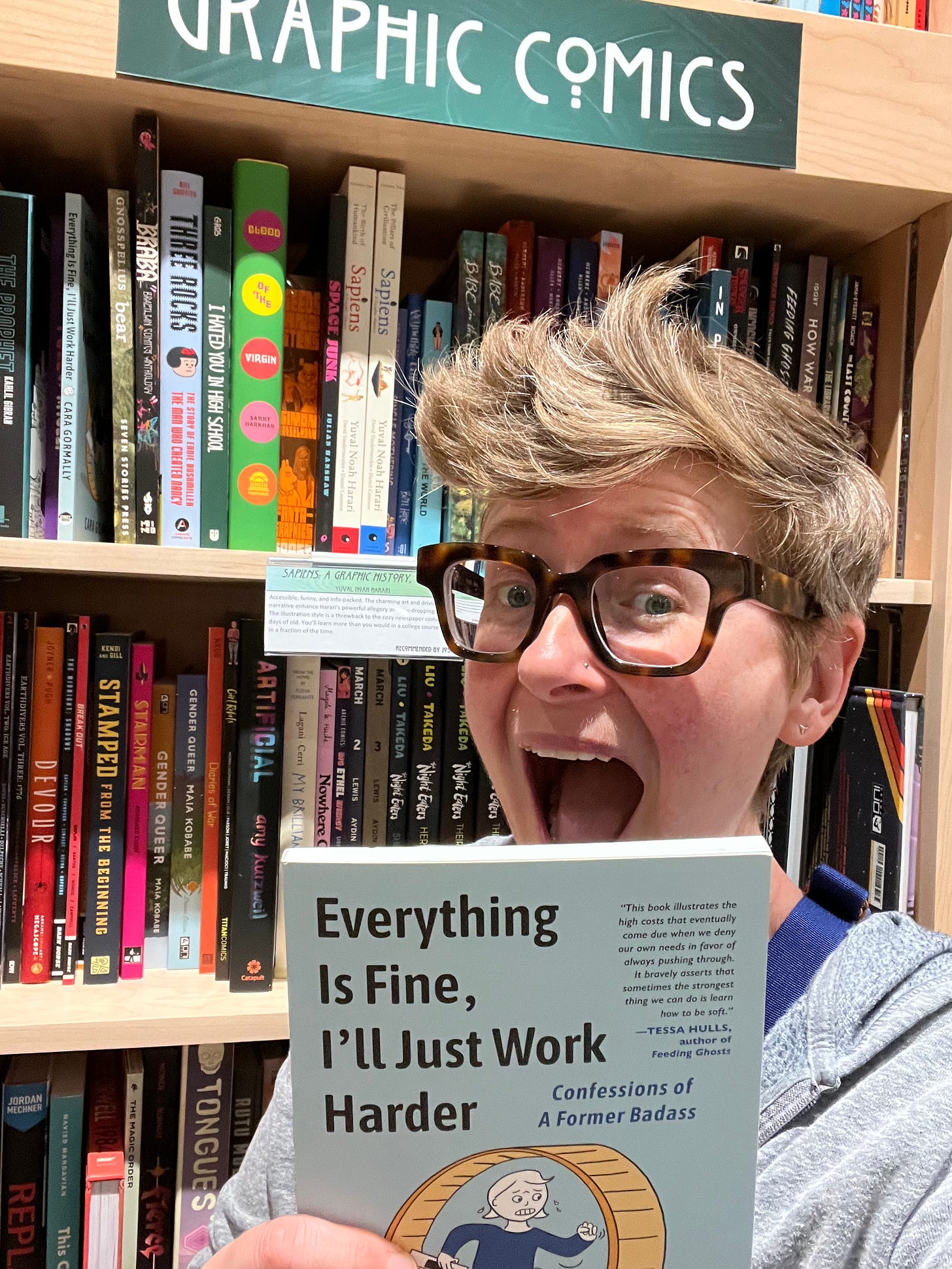 the author holding their book up with graphic novels on bookcases behind them