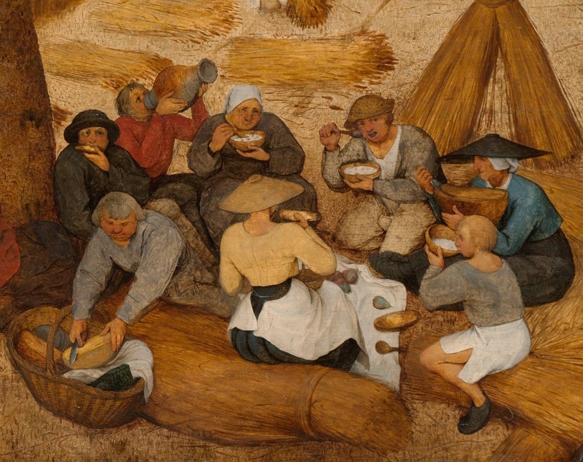 A group of men and women share a picnic lunch in the shade, a break from the harvest. A man slices bread and a woman slices cheese. There is butter nearby and fruit.