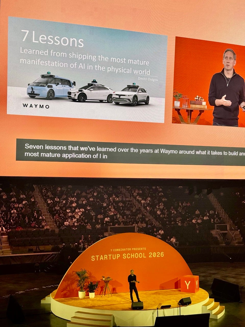 Conference-hall view of YC Startup School 2026: Dmitri Dolgov speaks on a round orange stage beneath a large screen showing a slide titled "7 Lessons - Learned from shipping the most mature manifestation of AI in the physical world," with three generations of Waymo vehicles pictured.