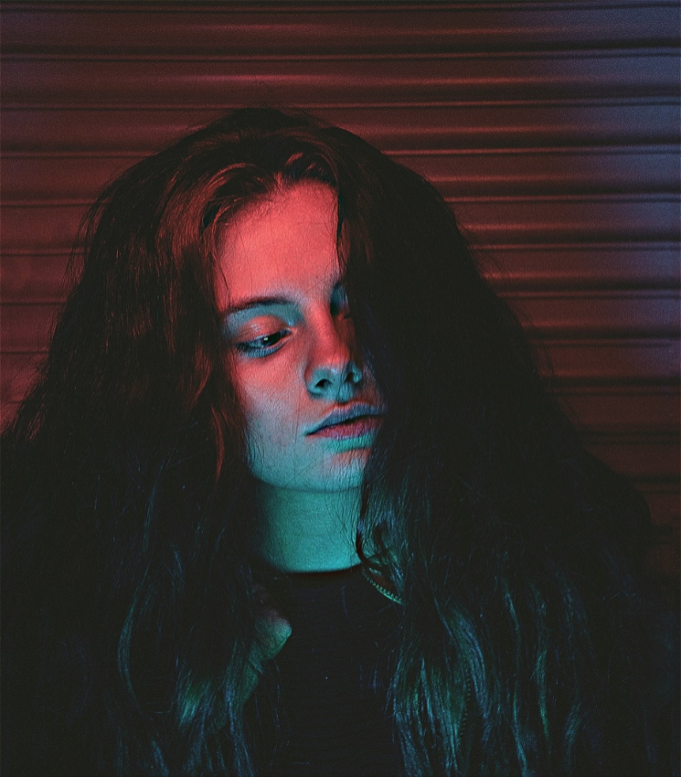 The head and shoulders of a young woman with long brunette hair and a black jacket and T-shirt, leaning against a grey rollerdoor. The lighting is a mainly dark but with a striking pink from above, and blue/green from below, subtly highlighting her face. She looks down, away from the camera, disinterested - or perhaps feigning it.